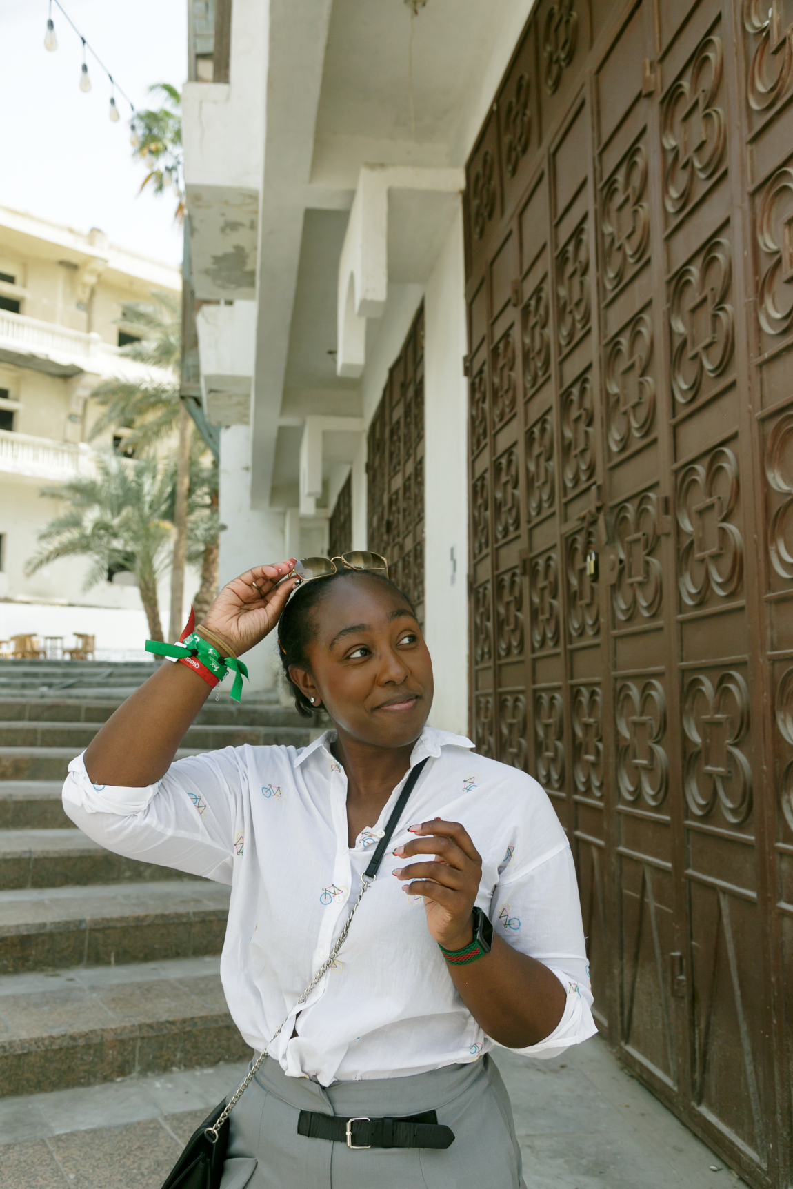 Young black woman wearing a button up shirt and crossbody bag, smiling to the left, standing at the bottom of a staircase in Jeddah, Saudi Arabia, while pulling her sunglasses off of her head