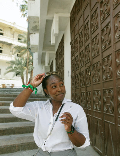 Young black woman wearing a button up shirt and crossbody bag, smiling to the left, standing at the bottom of a staircase in Jeddah, Saudi Arabia, while pulling her sunglasses off of her head