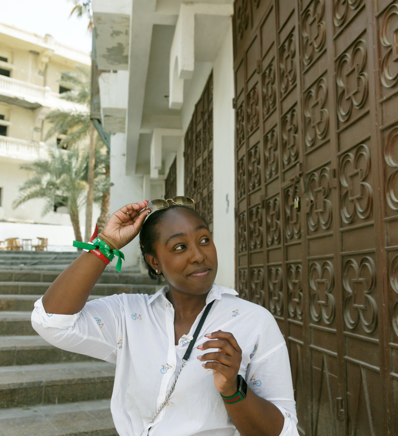 Young black woman wearing a button up shirt and crossbody bag, smiling to the left, standing at the bottom of a staircase in Jeddah, Saudi Arabia, while pulling her sunglasses off of her head