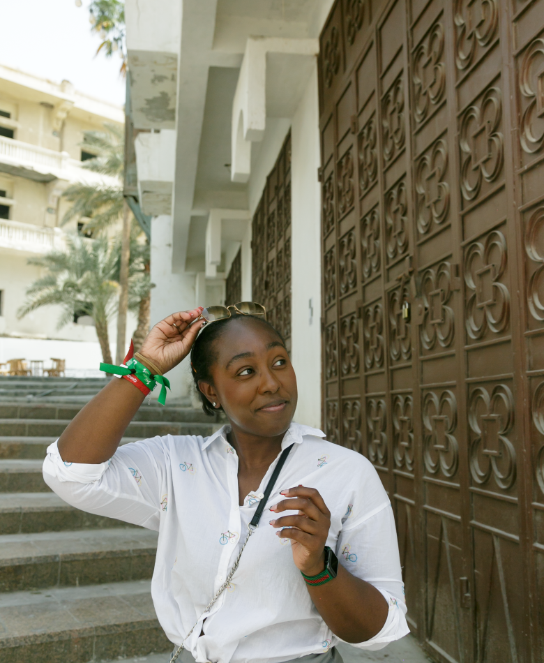Young black woman wearing a button up shirt and crossbody bag, smiling to the left, standing at the bottom of a staircase in Jeddah, Saudi Arabia, while pulling her sunglasses off of her head