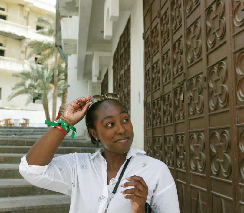 Young black woman wearing a button up shirt and crossbody bag, smiling to the left, standing at the bottom of a staircase in Jeddah, Saudi Arabia, while pulling her sunglasses off of her head